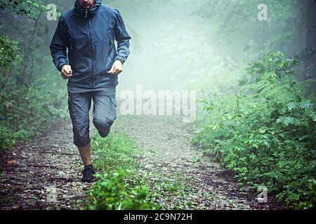 Primo piano di atleta maschile, indossando abiti all'aperto, che corre attraverso la foresta nebbiosa di mattina presto. Spazio per la copia disponibile. Foto Stock