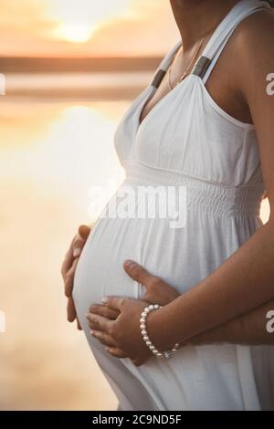 Primo piano ritratto di donna incinta felice in abito bianco insieme con il marito abbracciando al tramonto Foto Stock