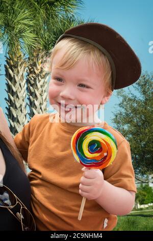 Felice ragazzo biondo di 2 anni con girl lollipop all'aperto in braccia di mamma Foto Stock