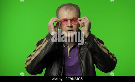 Il brutale e cool rocker man con giacca in pelle marrone indossa occhiali da sole rosa, guarda la fotocamera e sorride. Ritratto di ragazzo biker che posa su sfondo chiave chroma. Emozioni della gente. Spazio di copia Foto Stock