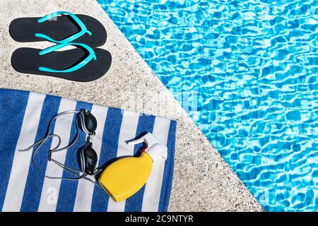 Infradito, crema solare e occhiali da bagno su un asciugamano a strisce blu e bianco sul bordo di una piscina. Spazio di copia. Vista dall'alto. Foto Stock