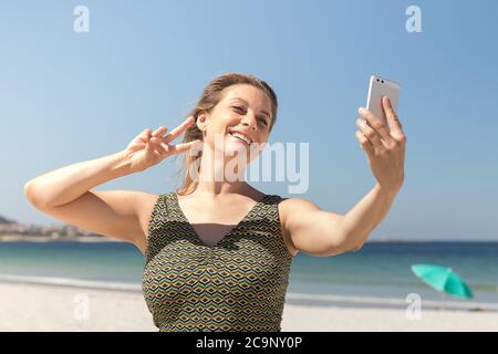 Primo piano di una donna bionda di mezza età in un abito verde che fa un selfie sulla spiaggia con il mare sullo sfondo Foto Stock