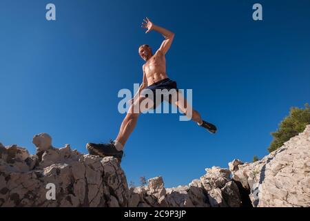 Attivo veloce corsa montagna sudato corpo runner muscolare che salta sopra la scogliera cleft durante il jogging mattutino. Attività sportive ad ampio angolo Foto Stock