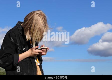 Ragazza teen che gioca online giochi da uno smartphone su sfondo cielo blu. Il concetto di dipendenza adolescente da un telefono cellulare e social network Foto Stock