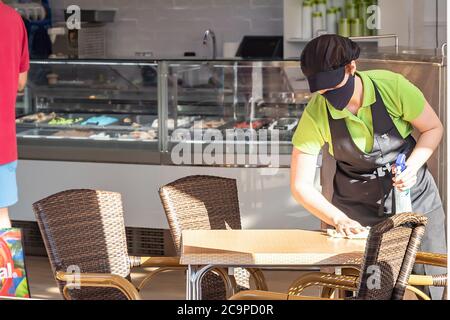 Huelva, Spagna - 4 luglio 2020: Una cameriera in maschera facciale sta pulendo un tavolo con liquido disinfettante nel centro commerciale Islantilla. Protezione ag Foto Stock