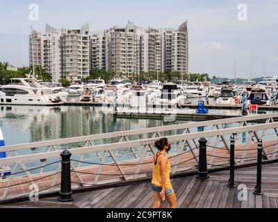 SINGAPORE – 31 LUGLIO 2020 – una donna asiatica cammina davanti a una fila di imbarcazioni da diporto ormeggiate sull'isola di Sentosa con condomini di lusso sullo sfondo Foto Stock