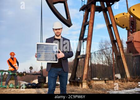 Uomo d'affari in giacca tuta, tenendo mini pannello solare, mostrando pollice in su. Operatore dell'olio che lavora con il martinetto della pompa sul campo dell'olio in background. Concetto di industria petrolifera e fonti energetiche alternative. Foto Stock