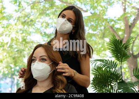 Elegante giovane parrucchiere con maschera per il viso forma i capelli con le mani ad un cliente rosso con capelli in un moderno salone di parrucchiere . Distanza sociale. Foto Stock