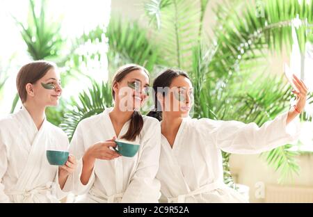 Compagnia di adorate amiche in accappatoi e con occhielli che prendono selfie sullo smartphone mentre si rilassano nel centro termale durante il fine settimana Foto Stock