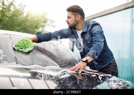 Vista orizzontale della pulizia dell'auto durante il servizio di autolavaggio all'aperto. Bell'uomo caucasico in abbigliamento casual che lava il parabrezza dell'auto con un mitten verde Foto Stock