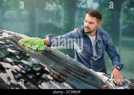 Vista orizzontale della pulizia dell'auto durante il servizio di autolavaggio all'aperto. Bell'uomo caucasico in abbigliamento casual che lava il parabrezza dell'auto con un mitten verde Foto Stock