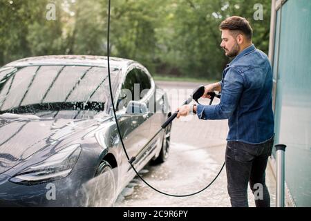 Caucasico giovane di stile messo a fuoco uomo in abiti casual lavando la sua moderna auto elettrica con una pistola ad acqua su auto self-service stazione di lavaggio all'aperto Foto Stock