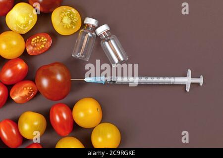 Concetto per alimenti geneticamente modificati con pomodori, siringa iniettata e fiale mediche vuote su sfondo marrone scuro Foto Stock