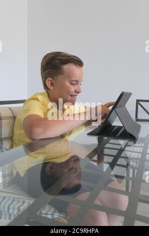 Ragazzo che parla con il compagno di classe durante la videochiamata sul tablet e discute di lavoro mentre si siede al tavolo in una stanza moderna e luminosa. Foto Stock