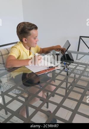 Ragazzo che parla con il compagno di classe durante la videochiamata sul tablet e discute di lavoro mentre si siede al tavolo in una stanza moderna e luminosa. Foto Stock