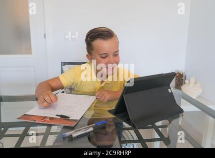 Ragazzo che parla con il compagno di classe durante la videochiamata sul tablet e discute di lavoro mentre si siede al tavolo in una stanza moderna e luminosa. Foto Stock