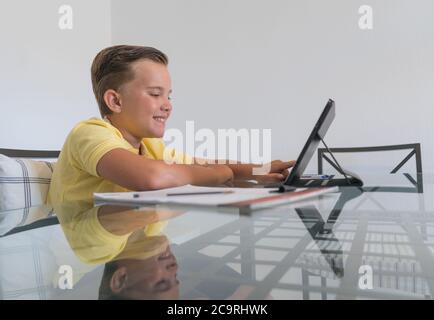 Ragazzo che parla con il compagno di classe durante la videochiamata sul tablet e discute di lavoro mentre si siede al tavolo in una stanza moderna e luminosa. Foto Stock