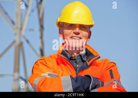 Ritratto di un ingegnere sorridente, un caposquadra o un lavoratore con indumenti protettivi e un elmetto di fronte a sfondo industriale con cielo blu Foto Stock