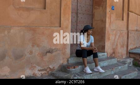 Donna seduta su gradini davanti alla porta con berretto e sneakers a Bologna Italia guardando verso sinistra Foto Stock