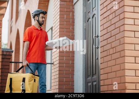 Il corriere consegna la pizza a porta. Giovane deliveryman con elmetto di sicurezza che regge una grande borsa gialla e una scatola Foto Stock