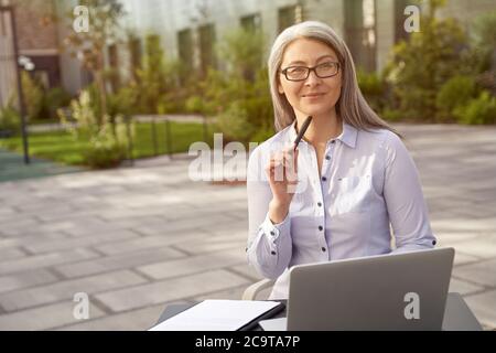 Pensare al business. Anziana bella donna d'affari che indossa occhiali seduti in bar all'aperto, lavorando su un computer portatile. Guardando da parte e sorridendo. Uomini d'affari, freelance Foto Stock