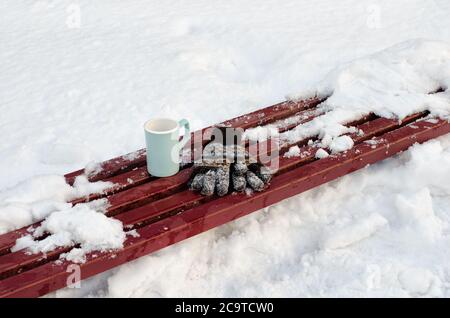 Guanti da donna e una tazza blu su una panchina tra la neve. Nessuno in una cornice Foto Stock