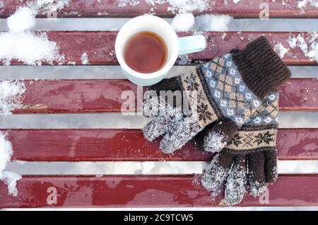 Tazza di tè e guanti in maglia da donna su una panca in inverno, la neve sotto. Vista dall'alto Foto Stock