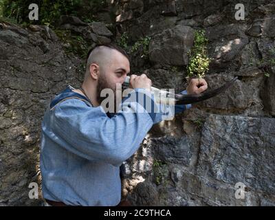 Un guerriero vichingo beve sidro da un corno appoggiato su un muro di pietra, immagine dell'uomo in abiti medievali Foto Stock