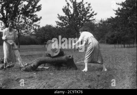 Anni '30, storico, due Signore che indossano abiti lunghi in un campo da qualche bosco, utilizzando una lunga mano troscatrice ha visto per tagliare un ceppo di albero, Inghilterra, Regno Unito. Le seghe a taglio trasversale o a taglio hanno un manico su ciascuna estremità e sono utilizzate da due persone per tagliare (perpendicolarmente) il grano di legno. Foto Stock