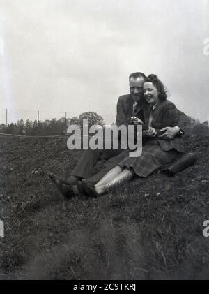 Anni '50, storico, formalmente vestito uomo in una tuta e una donna in una gonna e in cima, seduta su una riva erbosa ridendo insieme, con l'uomo abbracciando o tenendo la signora da vicino, Inghilterra, Regno Unito. Foto Stock