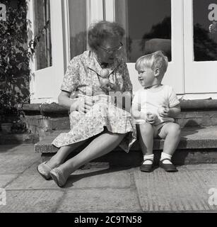 Anni '50, storico, 'è come lo fai'... una signora seduta con un ragazzo, suo nipote sui gradini alla porta posteriore di una casa che gli mostra come equilibrare due mele, mentre il ragazzino guarda tenendo le sue due mele in ogni mano, Inghilterra, Regno Unito. Foto Stock