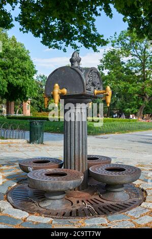 Vecchia fontana pubblica di ferro nel parco El Retiro a Madrid, Spagna. Quattro rubinetti. Lo sfondo un giorno di primavera con nuvole nel cielo e vegetazione verde. Foto Stock