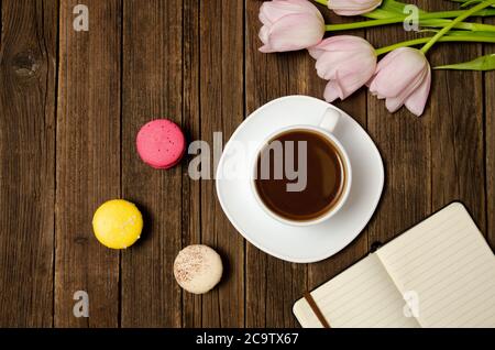 Tazza di caffè, macaron, tulipani rosa e taccuino su sfondo di legno. Vista dall'alto Foto Stock