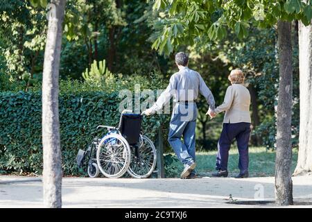 Madrid, Spagna - 7 luglio 2020: Una coppia anziana cammina di mano in mano attraverso il parco accanto a una sedia a rotelle. Foto Stock