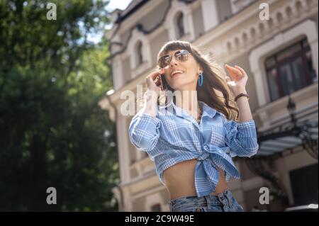 Gioiosa ragazza parla al telefono in occhiali da sole. Ritratto di una donna in occhiali da sole. Una ragazza giovane in una camicia blu plaid con gli occhiali parla al telefono Foto Stock