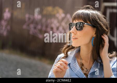 Ritratto esterno di yong bella donna sorridente felice che indossa occhiali da sole eleganti, blusa nera a pois, jeans blu mamma, con piccola borsa trapuntata. Mod Foto Stock