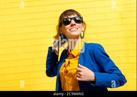 ragazza felice su uno sfondo giallo. Una ragazza in occhiali da sole, una camicia gialla e una giacca blu si contrappone al muro e sorride. Foto Stock