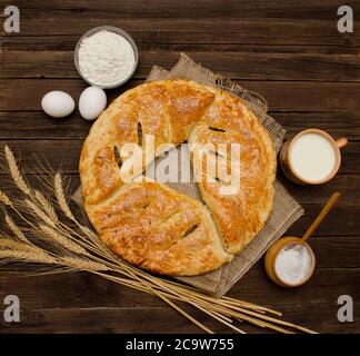 Torta su insaccatura, tazza di latte, ingredienti per la cottura, spighe di grano su fondo di legno. Foto Stock