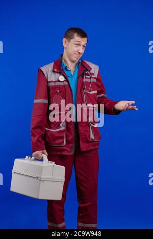 Ambulanza medico uomo in un vestito da lavoro e con un box in mano in posa su uno sfondo blu Foto Stock