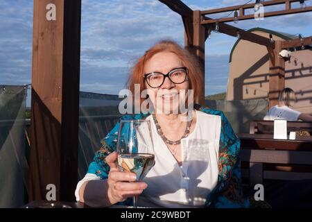 Una donna di età matura al tavolo di un ristorante in un'area aperta con un bicchiere di vino bianco secco Foto Stock