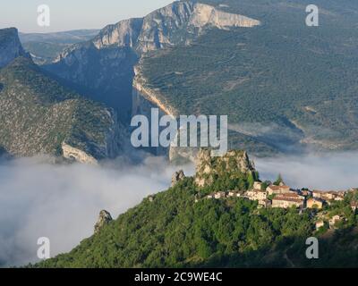 ARROCCATO VILLAGGIO DI ROUGON NELLA NEBBIA MATTUTINA CON LE MASSICCE SCOGLIERE DELLA GOLA DI VERDON PER SFONDO. Alpi dell'alta Provenza, Francia. Foto Stock