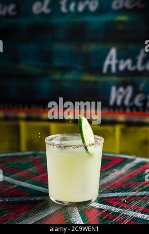 Una fetta di cetriolo che cade in un cocktail in un bicchiere a palla di roccia con un grande cubetto di ghiaccio, bordo speziato sul bicchiere Foto Stock