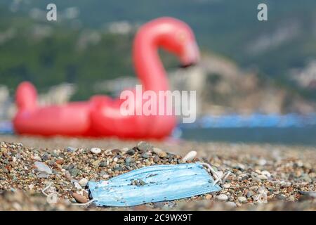 Becici, comune di Budva, Montenegro, Europa. 31 luglio 2020. BECICI E BUDVA, COMUNE DI BUDVA, MONTENEGRO - 31 LUGLIO 2020: Durante la pandemia COVID-19, le maschere mediche usate inquinano le spiagge europee, quindi queste maschere di fronte insieme con i detriti di plastica cadono nell'acqua, inquinando i mari e gli oceani. La maschera usata scartata si trova su una spiaggia di ciottoli, sullo sfondo un fenicottero rosa gonfiabile sulla riva. Credit: Andrey Nekrasov/ZUMA Wire/Alamy Live News Foto Stock