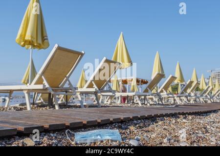 La maschera usata scartata si trova su una bellissima spiaggia di ciottoli, sedie a sdraio e ombrelloni sullo sfondo. Foto Stock