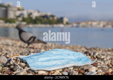 La maschera viso scartata usata si trova su una spiaggia di ciottoli, sullo sfondo una colomba passeggiate lungo la spiaggia. Foto Stock