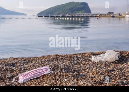 Scartato maschera usata e una borsa di plastica giace su una spiaggia di ciottoli, sullo sfondo il mare e l'isola Sveti Nikola. Foto Stock