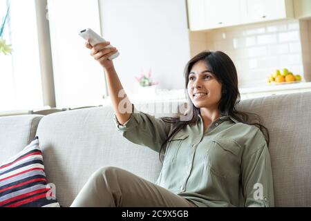 Donna indiana felice accende il sistema di aria condizionata che si rilassa sul divano a casa. Foto Stock