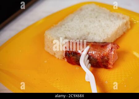 Salsiccia alla griglia su una forchetta e pane su un piatto di plastica giallo. Stoviglie monouso Foto Stock