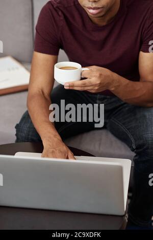 Immagine ritagliata di un giovane uomo nero che lavora su un computer portatile e beve una tazza di caffè Foto Stock