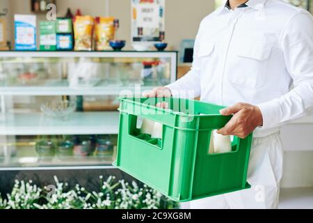 Immagine ritagliata dell'uomo da consegna in uniforme bianca che trasporta la cassa di plastica del latte al negozio di alimentari Foto Stock